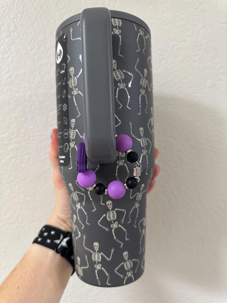 Puede incluir: Un vaso gris con asa, decorado con dise&ntilde;os de esqueletos plateados bailando. Un accesorio de cuentas moradas y negras cuelga del asa. El vaso lo sostiene una persona.