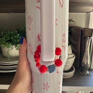 May include: White insulated tumbler with a handle, decorated with red seashell and bikini illustrations. A red and blue beaded bracelet is attached to the handle. The tumbler is held by a person.