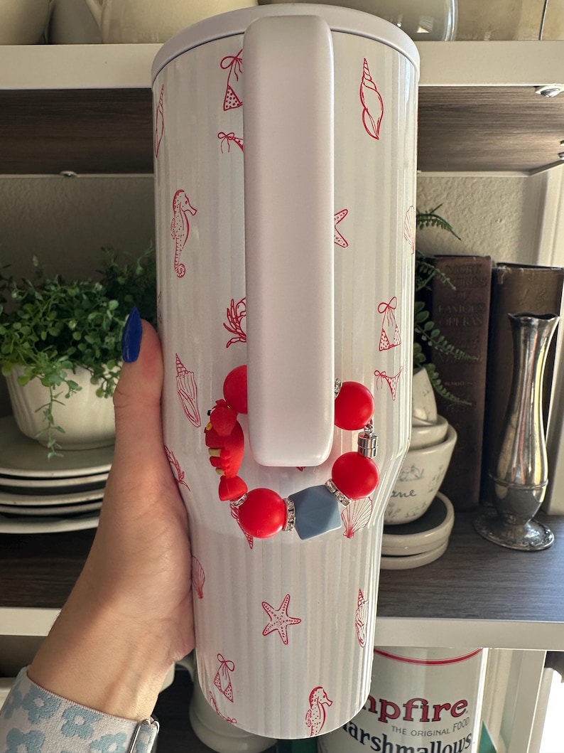 May include: White insulated tumbler with a handle, decorated with red seashell and bikini illustrations. A red and blue beaded bracelet is attached to the handle. The tumbler is held by a person.