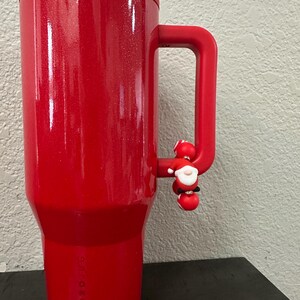 Może przedstawiać: Czerwony, izolowany kubek z uchwytem i małym urokiem Świętego Mikołaja. Kubek ma błyszczące wykończenie i pasującą czerwoną pokrywkę. Logo Hydro Jug jest widoczne z boku.