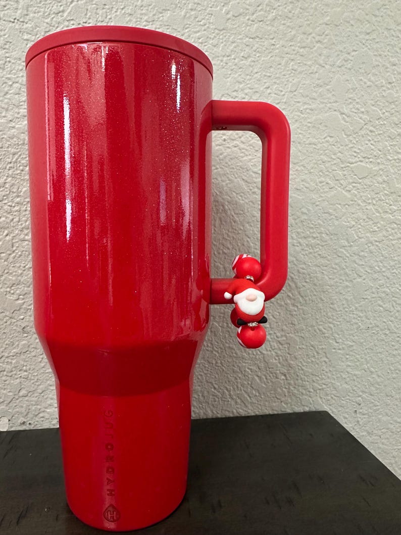 Może przedstawiać: Czerwony, izolowany kubek z uchwytem i małym urokiem Świętego Mikołaja. Kubek ma błyszczące wykończenie i pasującą czerwoną pokrywkę. Logo Hydro Jug jest widoczne z boku.