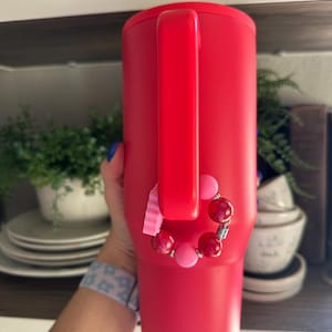 May include: A vibrant red insulated tumbler with a handle. A beaded bracelet with pink and red beads is attached to the handle. The tumbler is cylindrical and appears to be made of stainless steel.