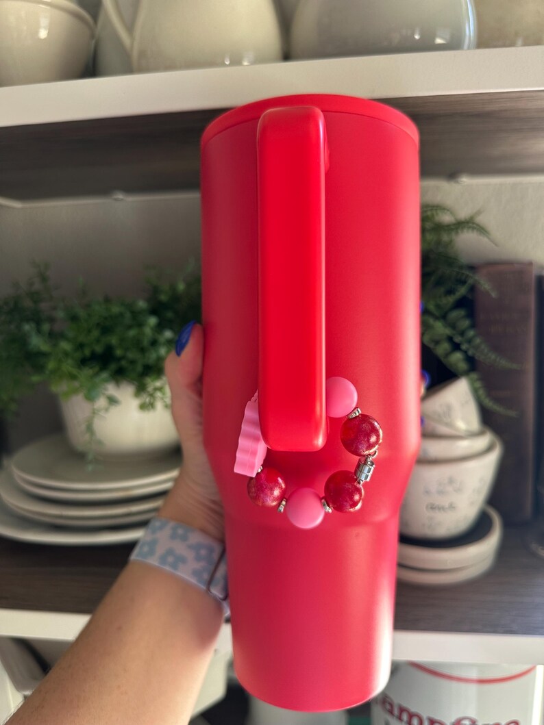 May include: A vibrant red insulated tumbler with a handle. A beaded bracelet with pink and red beads is attached to the handle. The tumbler is cylindrical and appears to be made of stainless steel.
