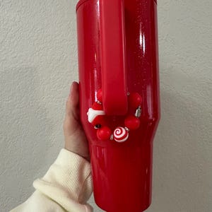 Może przedstawiać: Czerwony kubek termiczny z uchwytem i ozdobnym urokiem. Urok zawiera czerwone koraliki, laskę cukrową i figurkę Świętego Mikołaja. Kubek ma błyszczące wykończenie.