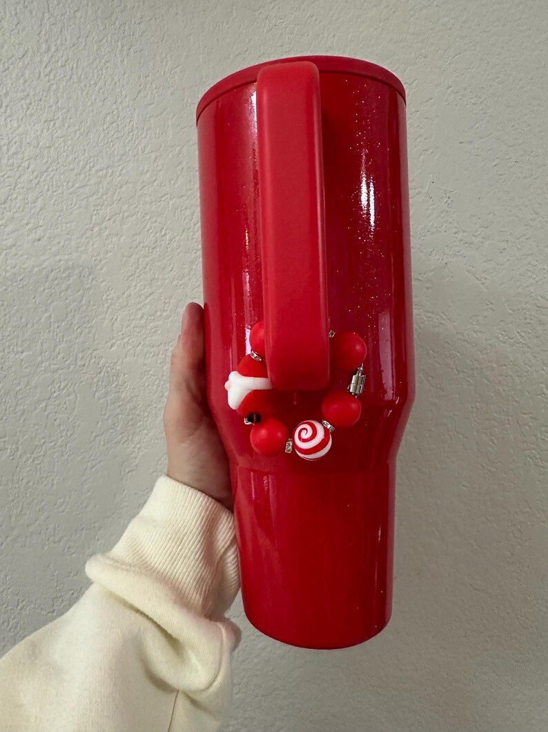 Może przedstawiać: Czerwony kubek termiczny z uchwytem i ozdobnym urokiem. Urok zawiera czerwone koraliki, laskę cukrową i figurkę Świętego Mikołaja. Kubek ma błyszczące wykończenie.