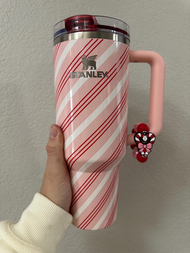 May include: A pink and white Stanley tumbler with a candy cane stripe design. The tumbler has a pink handle and a decorative charm. The Stanley logo is visible on the front of the tumbler.