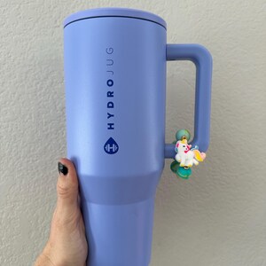 K&ouml;nnte beinhalten: Ein gro&szlig;er, lavendelfarbener Hydro Jug mit Griff und schwarzem Deckel. Auf der Seite des Jugs steht vertikal das Wort &bdquo;HYDRO JUG&ldquo; sowie ein Logo. Ein kleiner Einhorn-Anh&auml;nger ist am Griff befestigt.