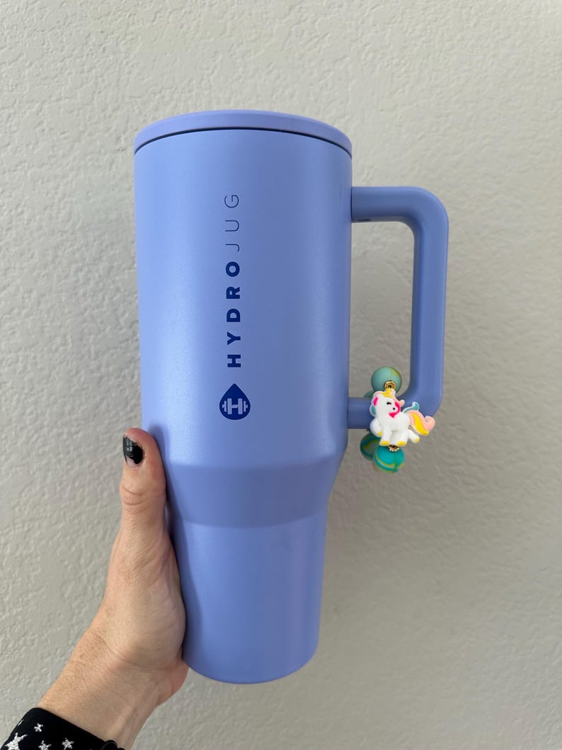 K&ouml;nnte beinhalten: Ein gro&szlig;er, lavendelfarbener Hydro Jug mit Griff und schwarzem Deckel. Auf der Seite des Jugs steht vertikal das Wort &bdquo;HYDRO JUG&ldquo; sowie ein Logo. Ein kleiner Einhorn-Anh&auml;nger ist am Griff befestigt.