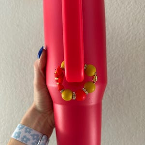 May include: A bright red insulated tumbler with a matching handle. A decorative beaded charm in yellow, red, and gold accents the tumbler. The tumbler is held by a person wearing a blue and white floral patterned watch.