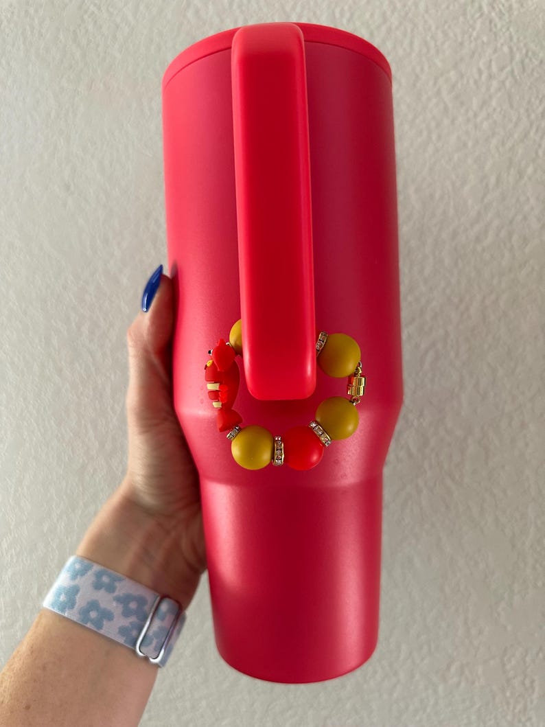 May include: A bright red insulated tumbler with a matching handle. A decorative beaded charm in yellow, red, and gold accents the tumbler. The tumbler is held by a person wearing a blue and white floral patterned watch.