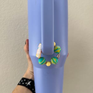 K&ouml;nnte beinhalten: Ein helllila isolierter Becher mit Henkel. Ein Perlen-Accessoire mit gelben, t&uuml;rkisfarbenen und wei&szlig;en Perlen ist am Henkel befestigt. Der Becher wird von einer Person gehalten.