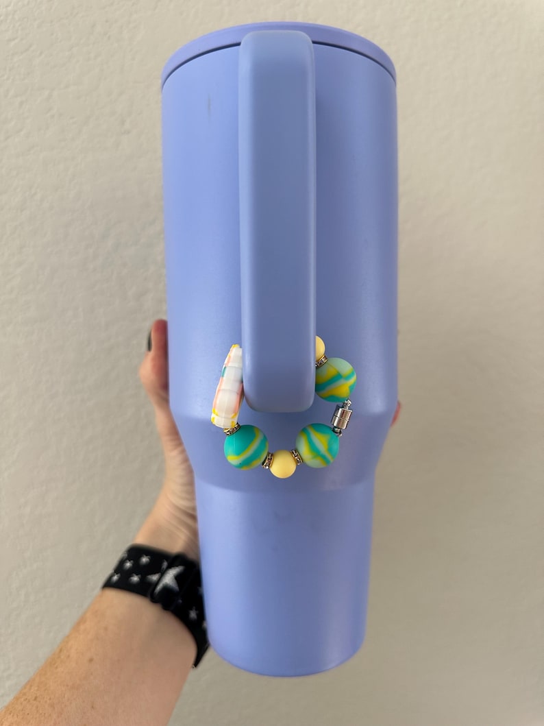 K&ouml;nnte beinhalten: Ein helllila isolierter Becher mit Henkel. Ein Perlen-Accessoire mit gelben, t&uuml;rkisfarbenen und wei&szlig;en Perlen ist am Henkel befestigt. Der Becher wird von einer Person gehalten.
