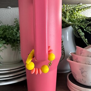 May include: A large, pink insulated tumbler with a handle. A decorative bracelet with yellow and orange beads is attached to the handle. The background includes white dishes and greenery.