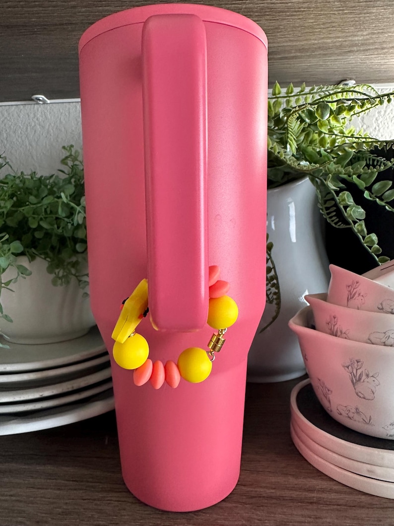 May include: A large, pink insulated tumbler with a handle. A decorative bracelet with yellow and orange beads is attached to the handle. The background includes white dishes and greenery.