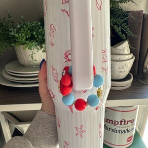 May include: White insulated tumbler with a handle, adorned with red seashell, bikini, and seahorse patterns. A beaded bracelet with red and blue beads is attached to the handle. The tumbler is approximately 10 inches tall.