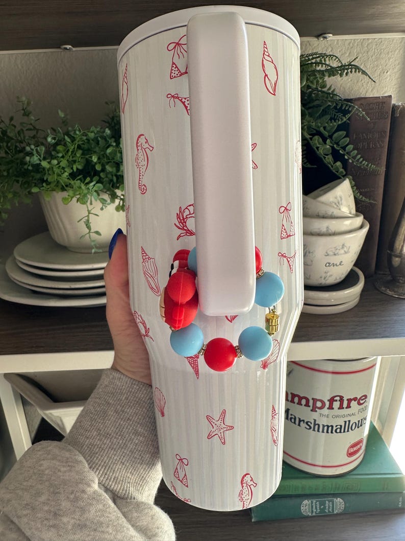 May include: White insulated tumbler with a handle, adorned with red seashell, bikini, and seahorse patterns. A beaded bracelet with red and blue beads is attached to the handle. The tumbler is approximately 10 inches tall.