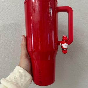 Może przedstawiać: Czerwony kubek termiczny z uchwytem i urokiem Świętego Mikołaja. Kubek ma błyszczące wykończenie i czarną pokrywkę. Uchwyt jest przymocowany do boku kubka. Zaprojektowany do utrzymywania zimnych lub gorących napoj&oacute;w.