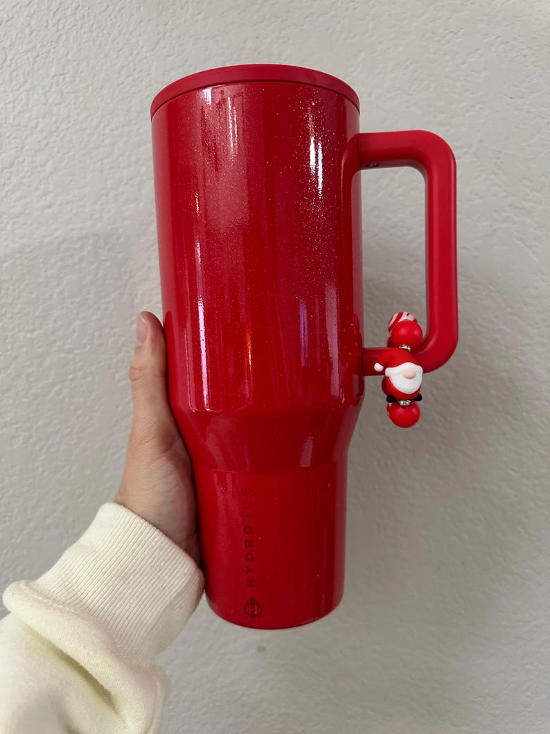 Może przedstawiać: Czerwony kubek termiczny z uchwytem i urokiem Świętego Mikołaja. Kubek ma błyszczące wykończenie i czarną pokrywkę. Uchwyt jest przymocowany do boku kubka. Zaprojektowany do utrzymywania zimnych lub gorących napoj&oacute;w.