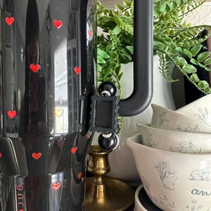 May include: Black Hydro Jug with red heart decals. The jug has a black handle and a black clip. The image also shows white bowls with a bunny and floral design, a brass candle holder, and a white vase with greenery.