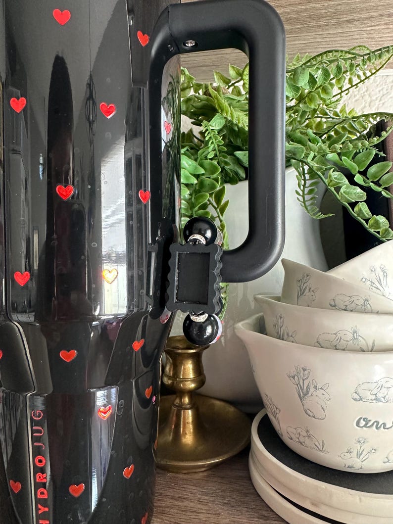 May include: Black Hydro Jug with red heart decals. The jug has a black handle and a black clip. The image also shows white bowls with a bunny and floral design, a brass candle holder, and a white vase with greenery.