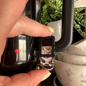 May include: A black coffee mug with a red heart design, held by a hand. A small, rectangular cat photo is attached to the mug. White bowls with rabbit illustrations are in the background.