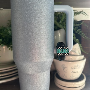 May include: A large, silver glitter tumbler with a light blue handle and rim. A small charm with the word "MAMA" in teal letters on a black and white checkered background is attached to the handle. The tumbler is displayed on a wooden shelf.