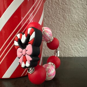 May include: A red and pink beaded bracelet with a candy cane charm. The charm is black, red, and white with a pink bow. The bracelet has red and pink beads with silver accents. The background includes a red and white striped container.