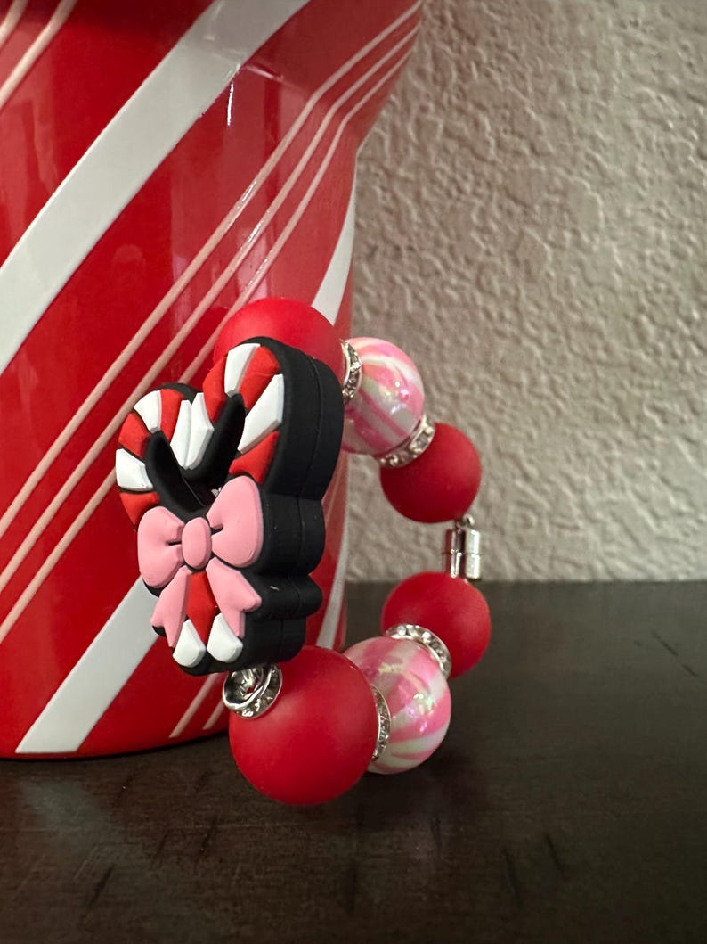 May include: A red and pink beaded bracelet with a candy cane charm. The charm is black, red, and white with a pink bow. The bracelet has red and pink beads with silver accents. The background includes a red and white striped container.