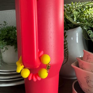 May include: A vibrant red insulated tumbler with a large handle. A yellow bracelet with yellow beads and a star charm is attached to the handle. The tumbler is on a wooden shelf with white dishes and greenery in the background.
