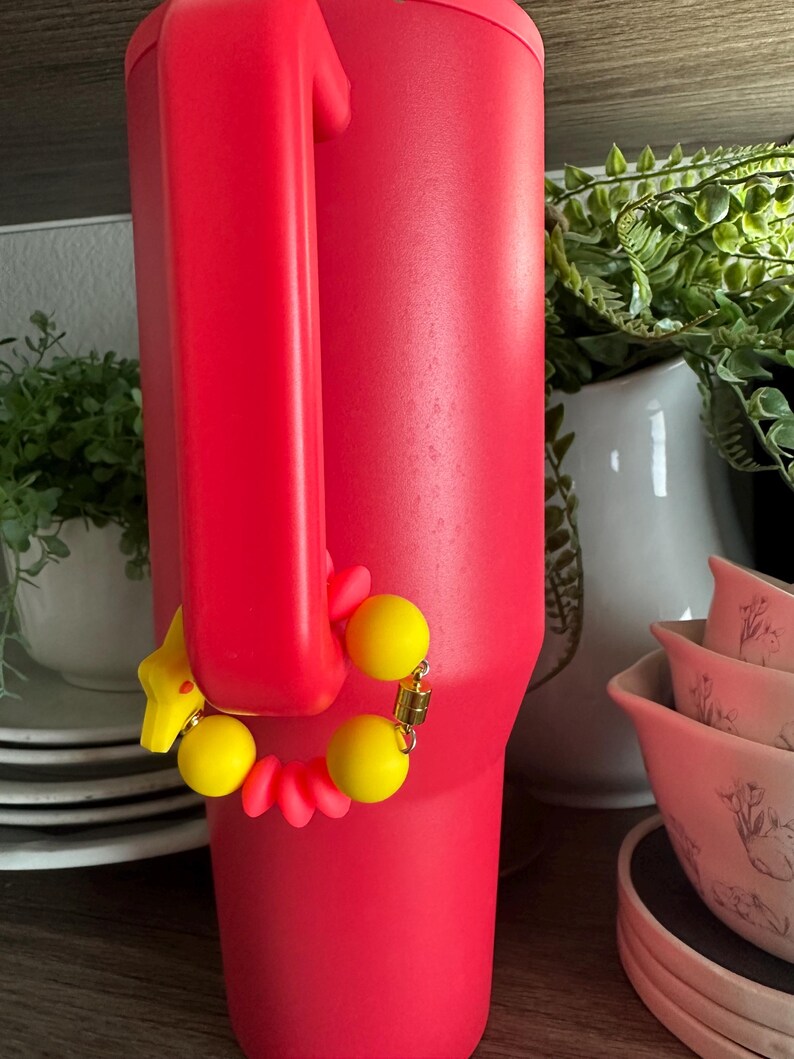 May include: A vibrant red insulated tumbler with a large handle. A yellow bracelet with yellow beads and a star charm is attached to the handle. The tumbler is on a wooden shelf with white dishes and greenery in the background.