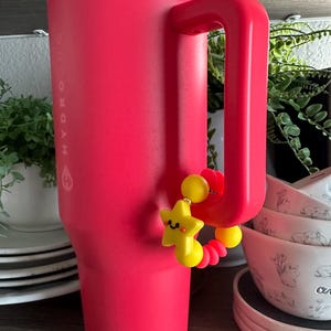 May include: A large, bright pink Hydrojug water bottle with a matching handle. A yellow star-shaped charm with a smiling face and red accents hangs from the handle. The bottle is on a wooden shelf with plates and bowls in the background.