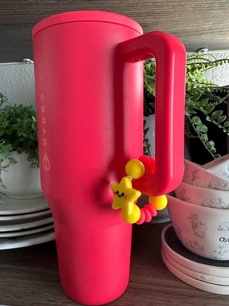 May include: A large, bright pink Hydrojug water bottle with a matching handle. A yellow star-shaped charm with a smiling face and red accents hangs from the handle. The bottle is on a wooden shelf with plates and bowls in the background.