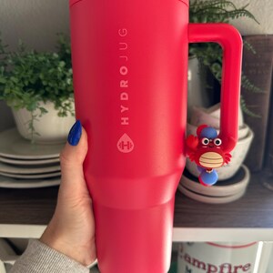 Puede incluir: Una botella de agua HydroJug de color rojo brillante con asa y un dije de cangrejo. La botella tiene el logotipo y el texto de HydroJug impresos. La botella es sostenida por una persona.