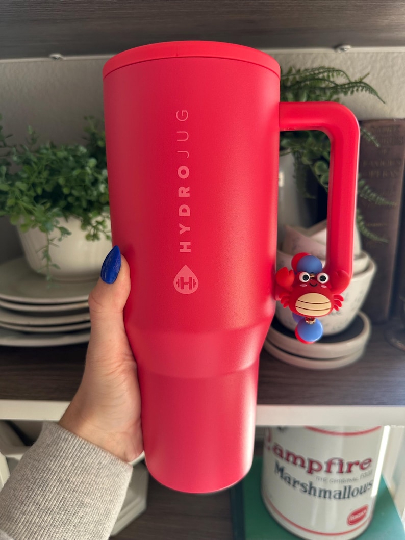 Puede incluir: Una botella de agua HydroJug de color rojo brillante con asa y un dije de cangrejo. La botella tiene el logotipo y el texto de HydroJug impresos. La botella es sostenida por una persona.