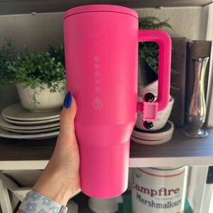 Pode incluir: Um copo Hydrojug rosa brilhante com uma al&ccedil;a. O copo tem o nome da marca impresso verticalmente na lateral. A al&ccedil;a &eacute; fixada com uma pequena pe&ccedil;a articulada rosa. O copo &eacute; segurado por uma pessoa com esmalte de unha azul e uma pulseira com estampa floral.