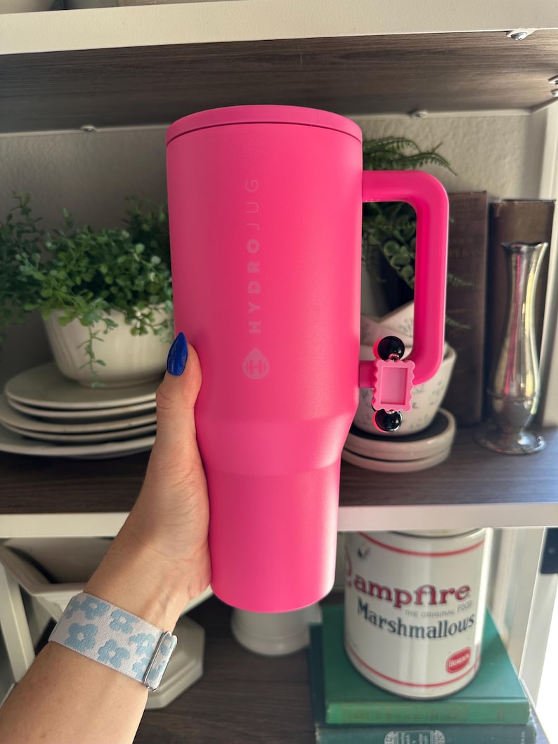 Pode incluir: Um copo Hydrojug rosa brilhante com uma al&ccedil;a. O copo tem o nome da marca impresso verticalmente na lateral. A al&ccedil;a &eacute; fixada com uma pequena pe&ccedil;a articulada rosa. O copo &eacute; segurado por uma pessoa com esmalte de unha azul e uma pulseira com estampa floral.