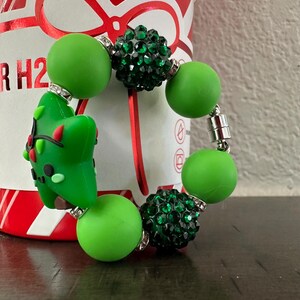 Pode incluir: Uma pulseira de contas verdes com um berloque de &aacute;rvore de Natal. A pulseira apresenta contas verdes grandes e redondas e contas verdes menores e brilhantes. O berloque da &aacute;rvore de Natal &eacute; verde com detalhes em vermelho e preto. A pulseira &eacute; presa com um fecho magn&eacute;tico.