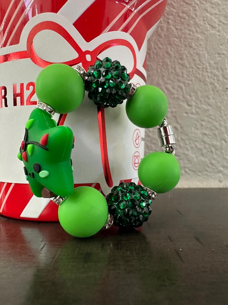 Pode incluir: Uma pulseira de contas verdes com um berloque de &aacute;rvore de Natal. A pulseira apresenta contas verdes grandes e redondas e contas verdes menores e brilhantes. O berloque da &aacute;rvore de Natal &eacute; verde com detalhes em vermelho e preto. A pulseira &eacute; presa com um fecho magn&eacute;tico.