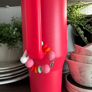 May include: A large, bright red insulated tumbler with a handle. A beaded charm bracelet in shades of pink, orange, and turquoise is attached to the handle. The tumbler is on a wooden shelf with other items in the background.