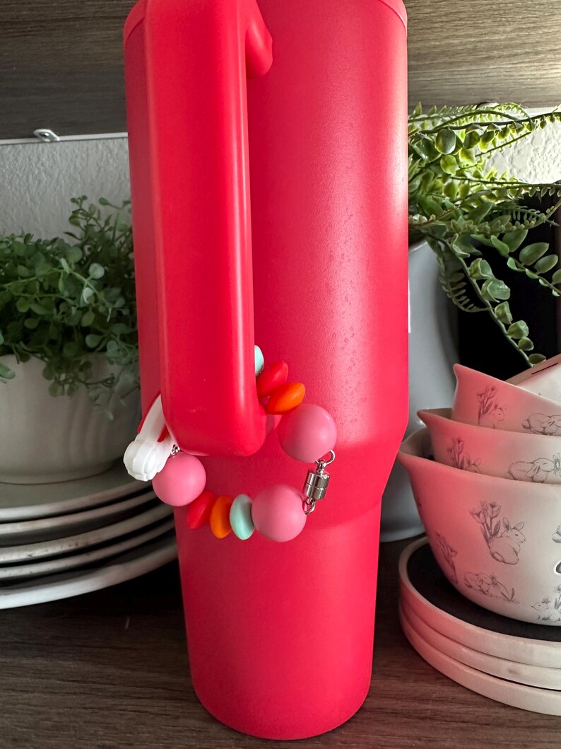 May include: A large, bright red insulated tumbler with a handle. A beaded charm bracelet in shades of pink, orange, and turquoise is attached to the handle. The tumbler is on a wooden shelf with other items in the background.