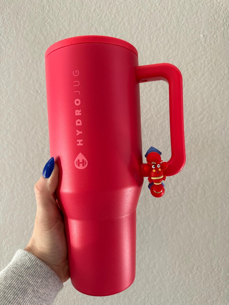 以下が含まれることがあります： ハンドルとロブスターの装飾が付いた赤いHydrojugウォーターボトル。「HYDROJUG」の文字が縦にプリントされています。円筒形のボトルには赤い蓋が付いています。ハンドルは側面に付いています。