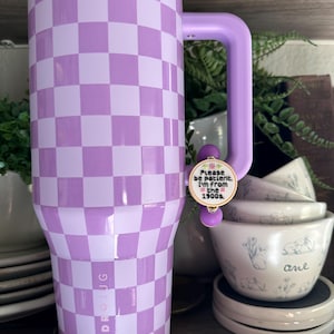 Puede incluir: Un vaso grande y aislado con un patr&oacute;n de cuadros lavanda y blanco. El vaso tiene un asa y una tapa a juego. Un peque&ntilde;o dije con el texto "Please be patient. I'm from the 1900s" est&aacute; adjunto al asa. La marca "HYDROJUG" est&aacute; impresa en el lateral.