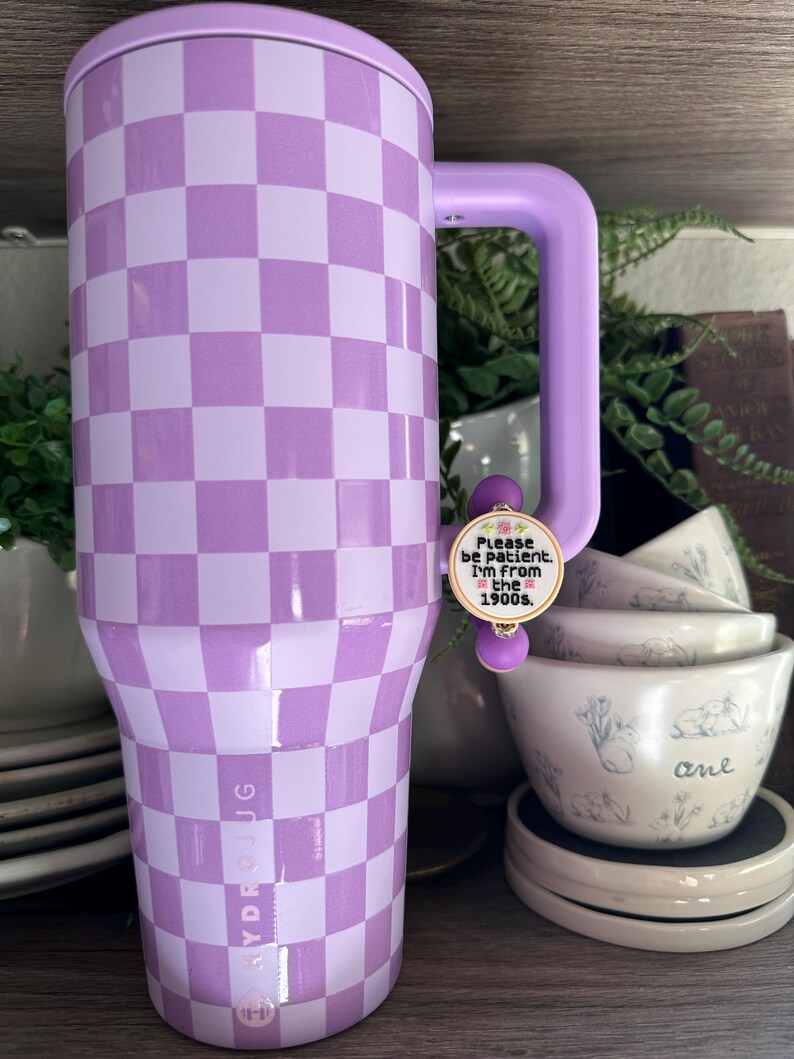 Puede incluir: Un vaso grande y aislado con un patr&oacute;n de cuadros lavanda y blanco. El vaso tiene un asa y una tapa a juego. Un peque&ntilde;o dije con el texto "Please be patient. I'm from the 1900s" est&aacute; adjunto al asa. La marca "HYDROJUG" est&aacute; impresa en el lateral.