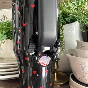 May include: Black tumbler with red heart accents. The tumbler has a black handle and a beaded bracelet with black beads and a clear bead with red confetti. The tumbler is decorated with small red hearts.