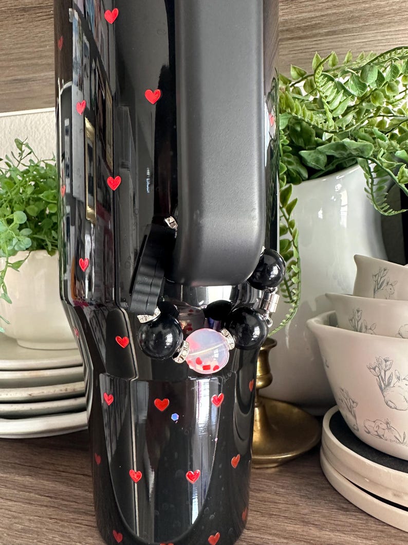 May include: Black tumbler with red heart accents. The tumbler has a black handle and a beaded bracelet with black beads and a clear bead with red confetti. The tumbler is decorated with small red hearts.