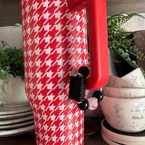 May include: A red and white houndstooth pattern tumbler with a red handle and lid. A black and clear beaded charm is attached to the handle. The tumbler is displayed on a shelf with other kitchenware.