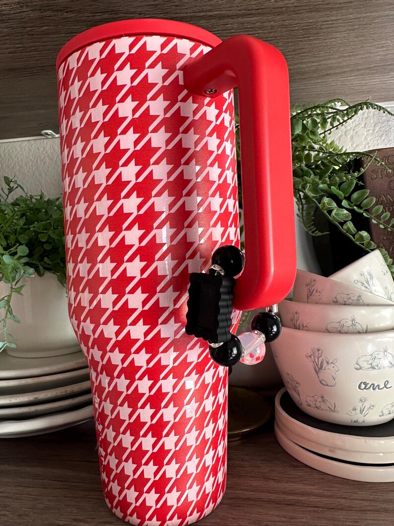 May include: A red and white houndstooth pattern tumbler with a red handle and lid. A black and clear beaded charm is attached to the handle. The tumbler is displayed on a shelf with other kitchenware.
