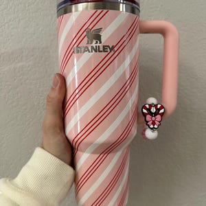 May include: A pink and white striped Stanley tumbler with a red and pink candy cane charm. The tumbler has a handle and a clear lid. The Stanley logo is visible on the side. The tumbler is approximately 10 inches tall.