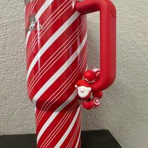 Może przedstawiać: Czerwono-biały kubek Stanley w paski z czerwonym uchwytem i zawieszką Świętego Mikołaja. Kubek ma obręcz ze stali nierdzewnej i przezroczystą pokrywkę. Logo Stanley jest widoczne z boku.