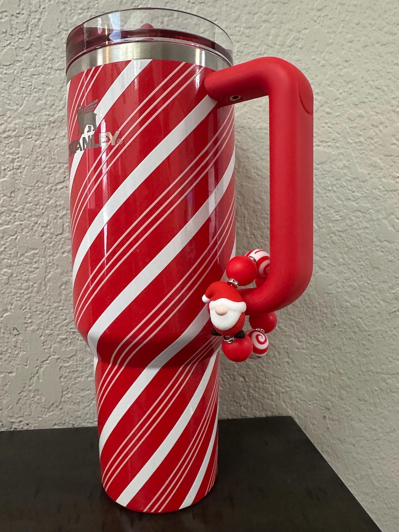 Może przedstawiać: Czerwono-biały kubek Stanley w paski z czerwonym uchwytem i zawieszką Świętego Mikołaja. Kubek ma obręcz ze stali nierdzewnej i przezroczystą pokrywkę. Logo Stanley jest widoczne z boku.